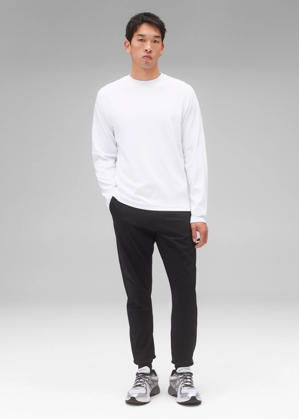 A man modeling a white long-sleeve crewneck shirt paired with black jogger pants and gray athletic sneakers, standing against a neutral gray studio background.