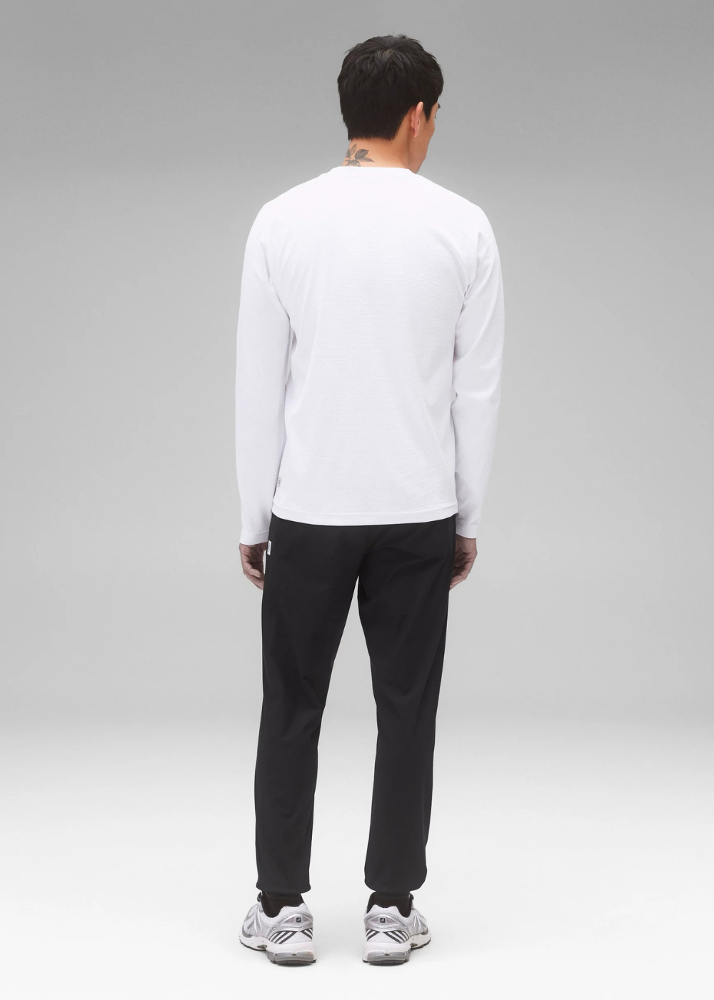 A man viewed from behind wearing a white long-sleeve crewneck shirt, black pants, and white sneakers, standing against a light gray studio background.