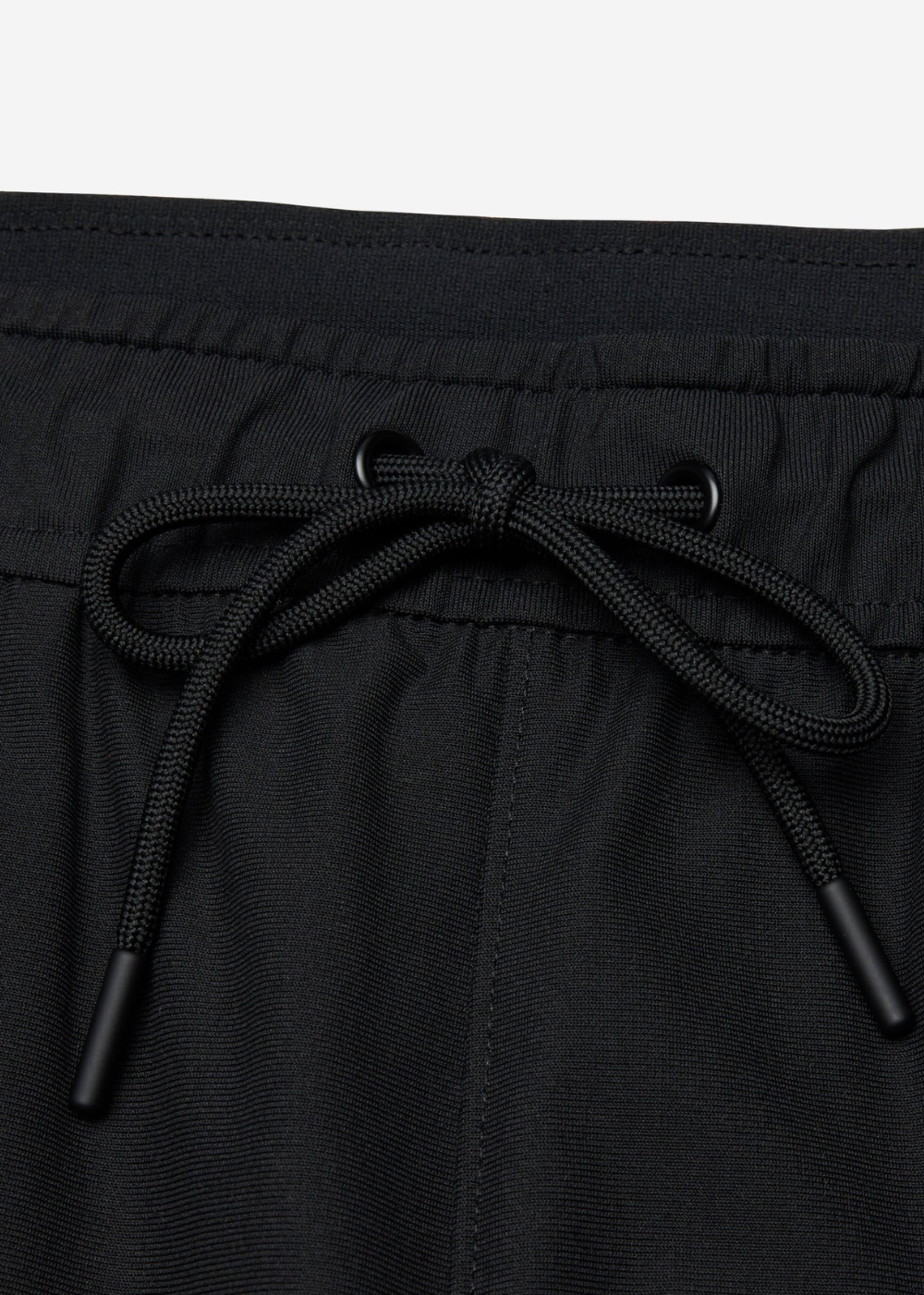 Close-up of black drawstring shorts showing the waistband, black drawstring tied in a bow, and black plastic tip ends against a light gray background.