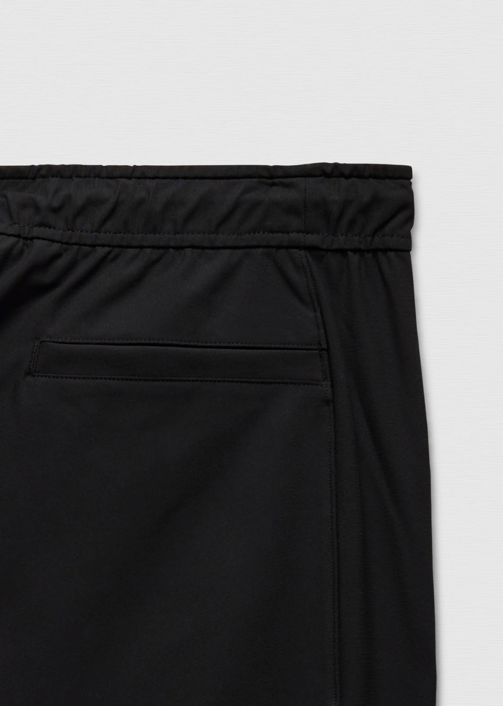 Close-up of black trousers showing the elastic waistband and a side pocket, photographed against a light gray background.