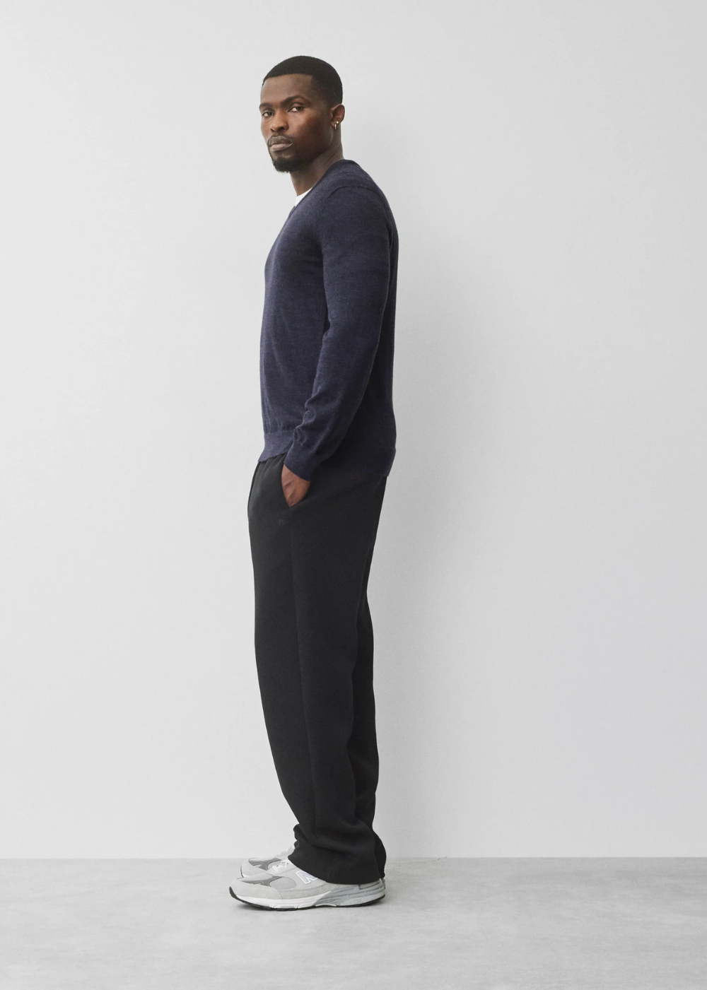 A man in a side profile wearing a dark navy long-sleeve knit sweater, black sweatpants, and white sneakers, standing against a light gray studio background.
