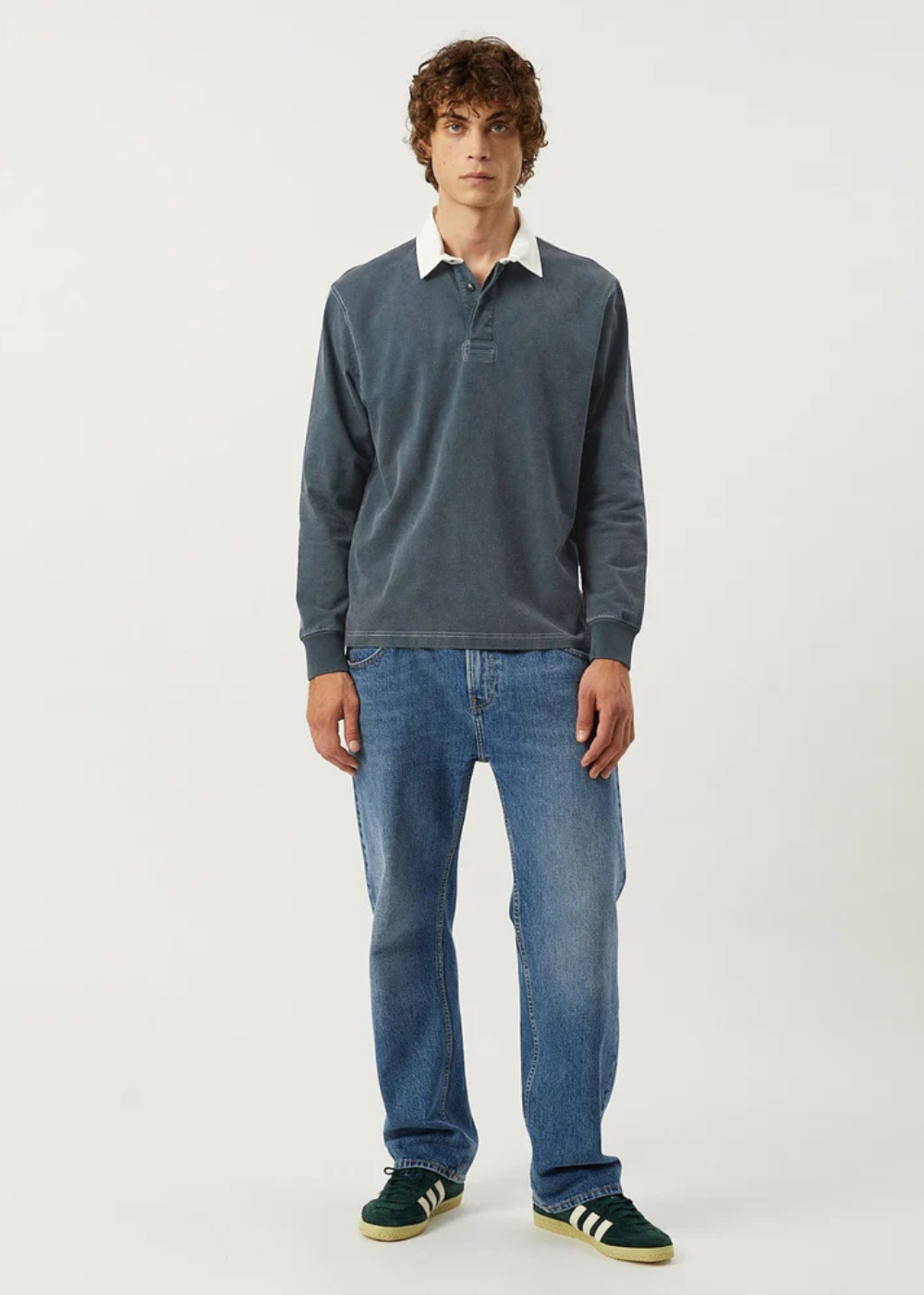 A man modeling a slate blue long-sleeve polo shirt with a white collar, paired with blue jeans and dark green sneakers, against a plain white studio background.