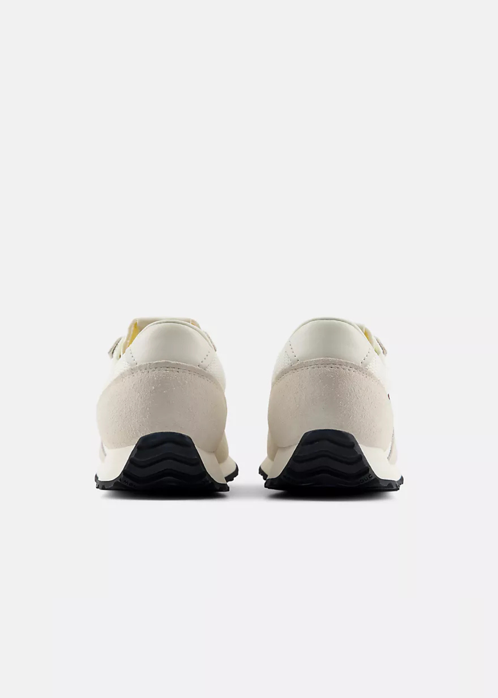 A pair of low-top sneakers shown from the back, featuring off-white leather uppers and black wavy rubber soles, set against a plain light gray background.