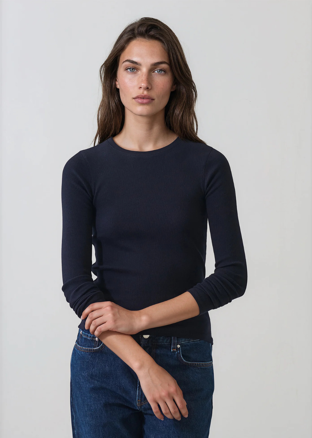 A woman modeling a dark navy blue long-sleeve crewneck sweater paired with blue jeans, standing against a light gray studio background.