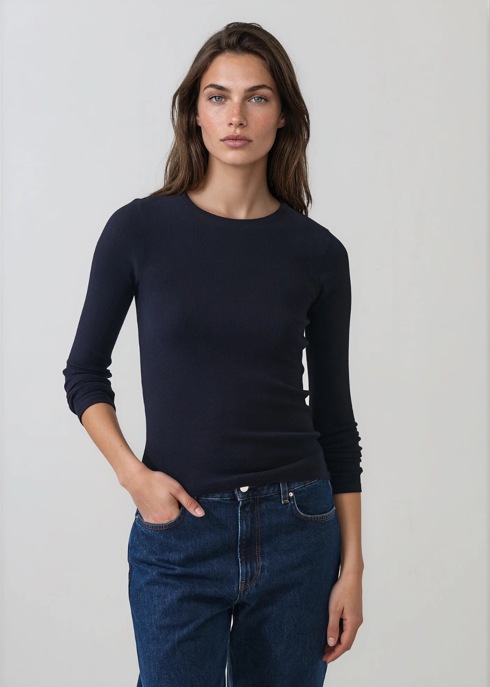 A woman modeling a dark navy long-sleeve crewneck top paired with blue jeans, standing against a light gray studio background.