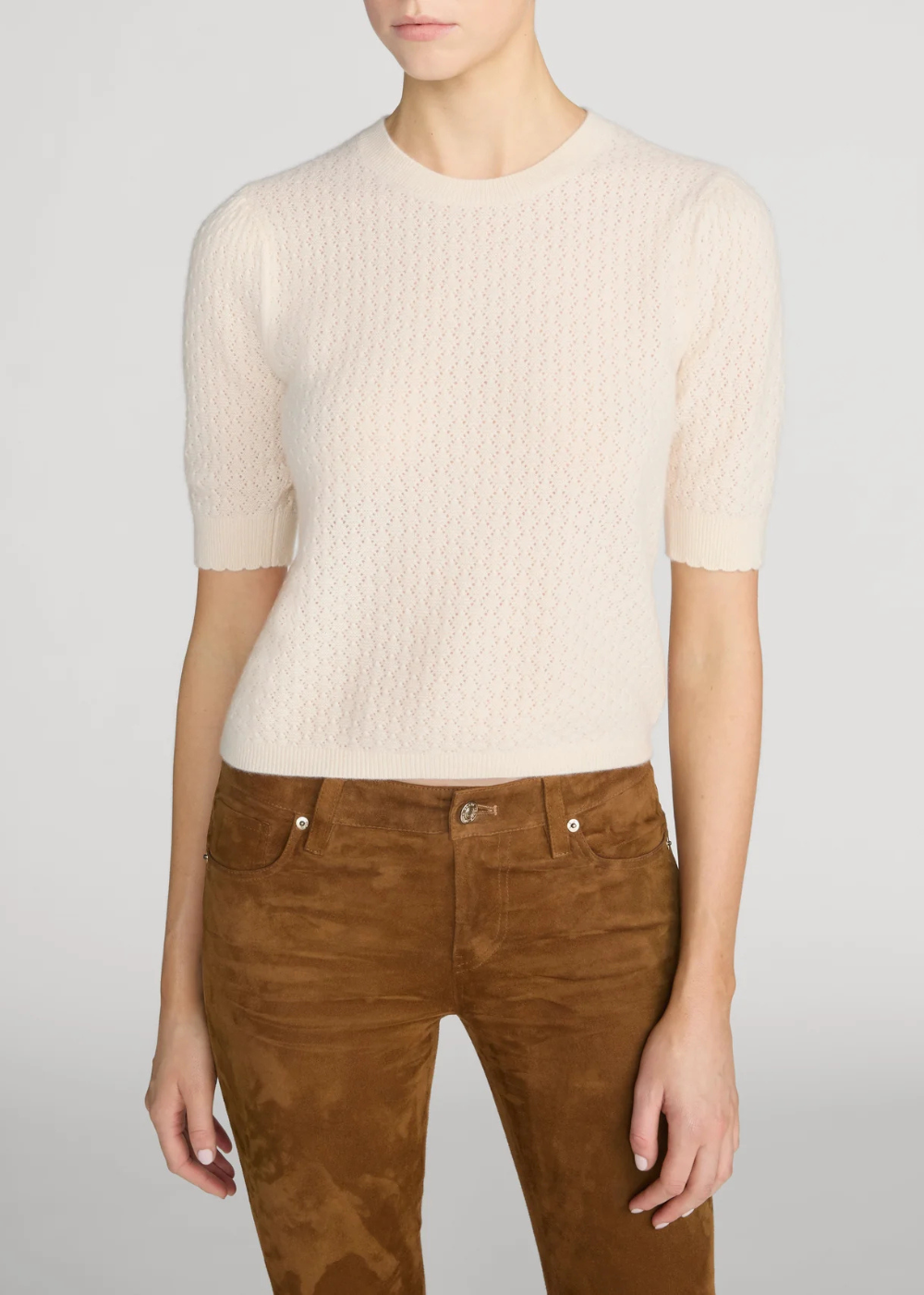 A model wearing a cream-colored short-sleeve knit sweater with a textured cable pattern, paired with brown suede pants, against a light gray studio background.