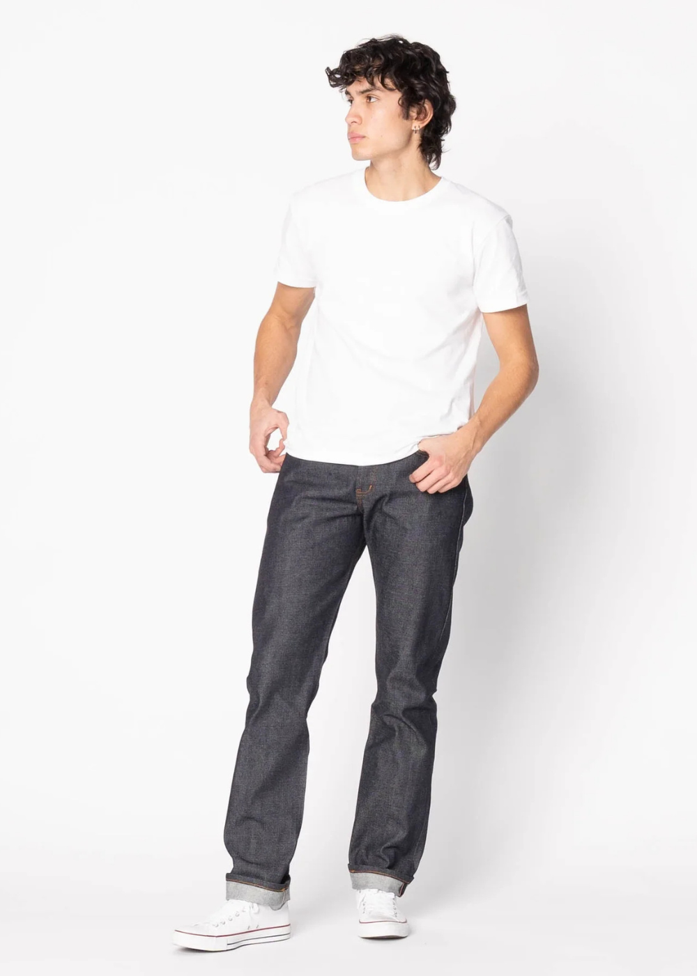 A man wearing a plain white crew-neck t-shirt, dark indigo slim-fit jeans, and white sneakers, standing against a solid white studio background.