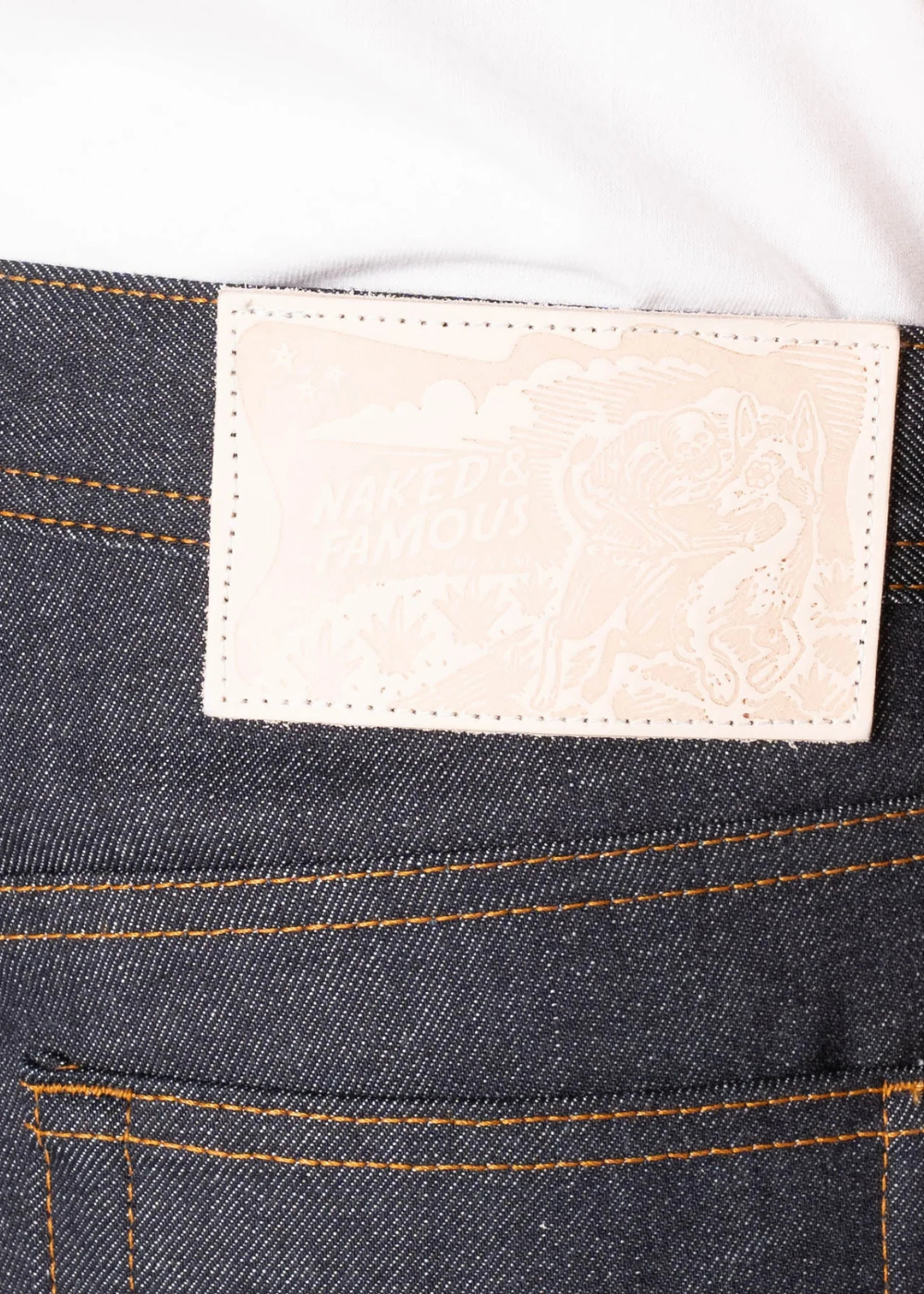 Close-up of the back waistband of dark indigo denim jeans with orange contrast stitching, featuring a cream-colored leather patch with a subtle embossed design.