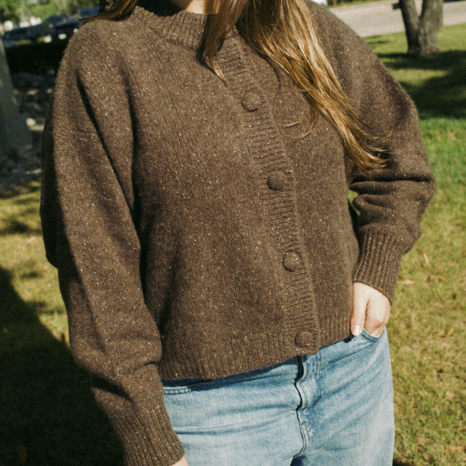 Women model wearing a brown cardigan sweater with blue denim. Photo is taken outside against trees and grass.