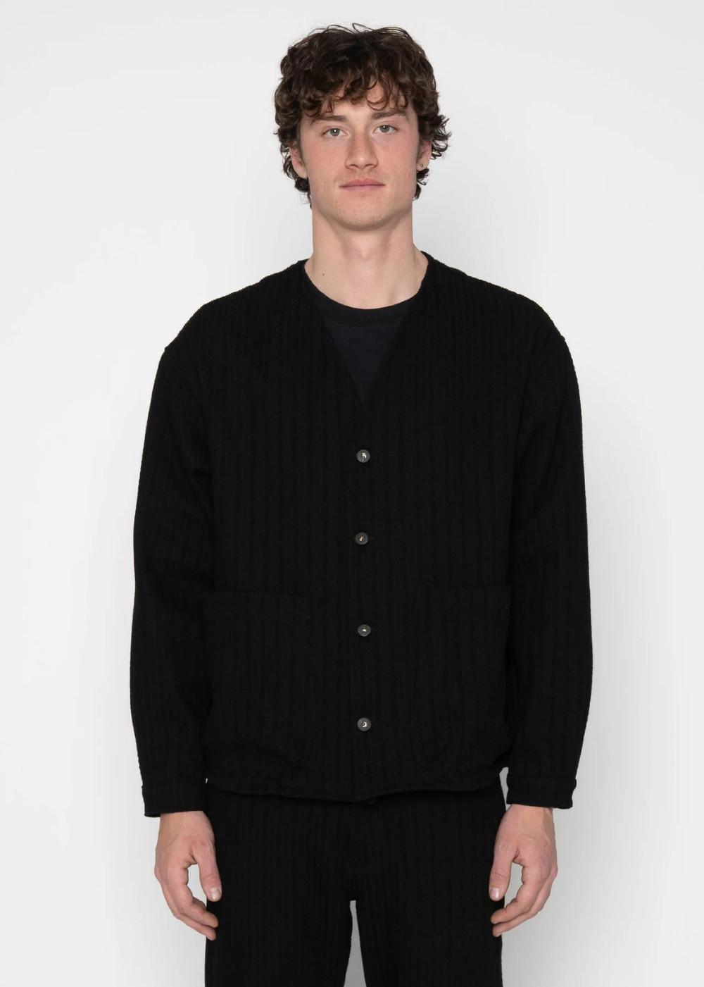 A man wearing a black ribbed knit cardigan over a black t-shirt and black pants, standing against a light gray studio background.