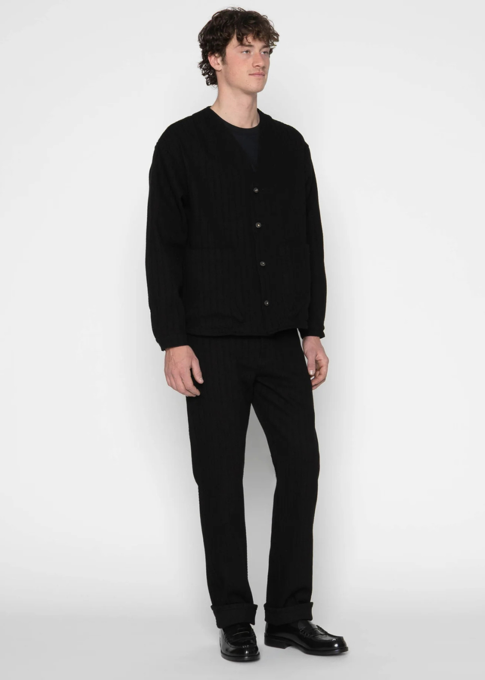 A man modeling a black ribbed knit cardigan sweater, black trousers, and black leather shoes against a plain light gray studio background.