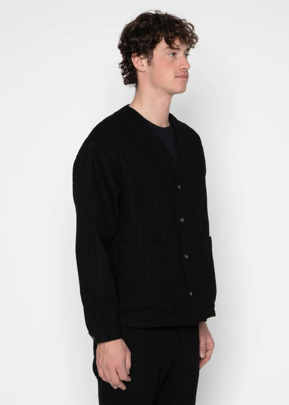 A man wearing a black ribbed knit cardigan over a black t-shirt and black trousers, standing in profile against a plain white studio background.