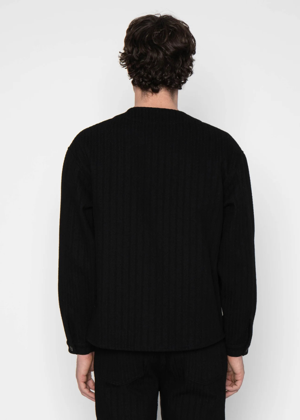 A man viewed from behind wearing a black ribbed long-sleeve top and black pants against a plain light gray studio background.