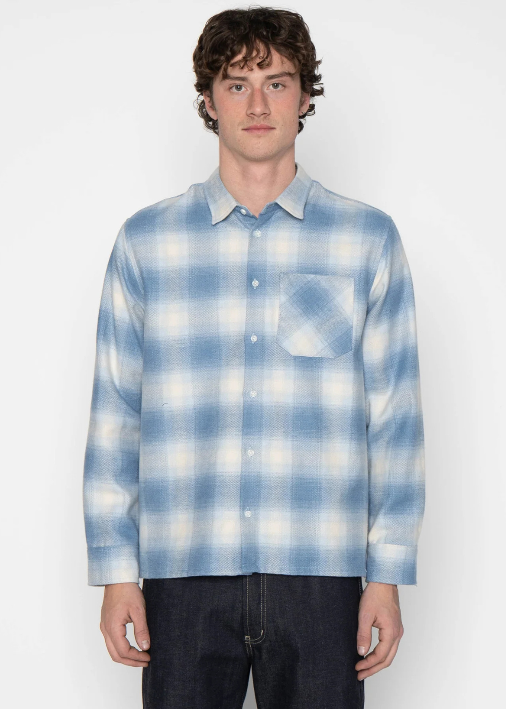 A man wearing a light blue and white plaid long-sleeve button-up shirt with a chest pocket, paired with dark jeans, standing against a plain light gray background.