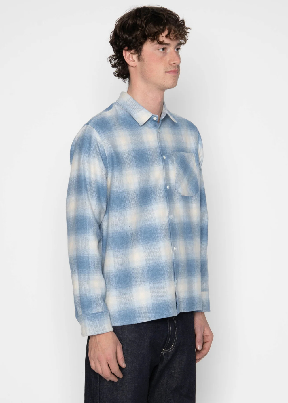 A man wearing a light blue and white plaid button-down shirt, paired with dark jeans, standing against a plain white studio background.