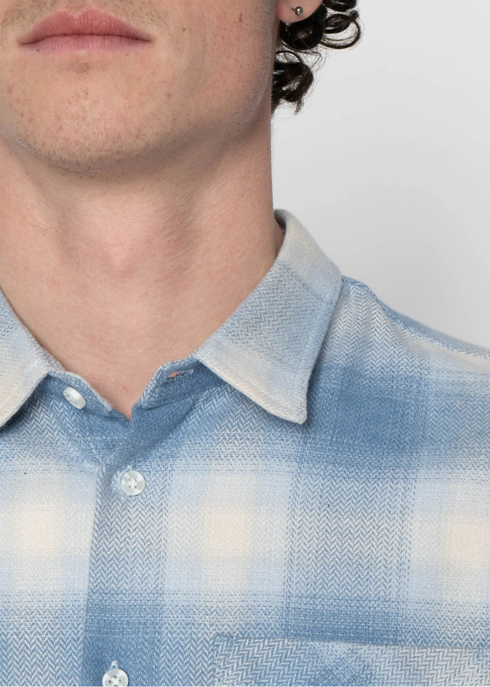 Close-up of a person wearing a light blue and white plaid button-down shirt, showing the collar, buttons, and part of the chest against a plain light background.