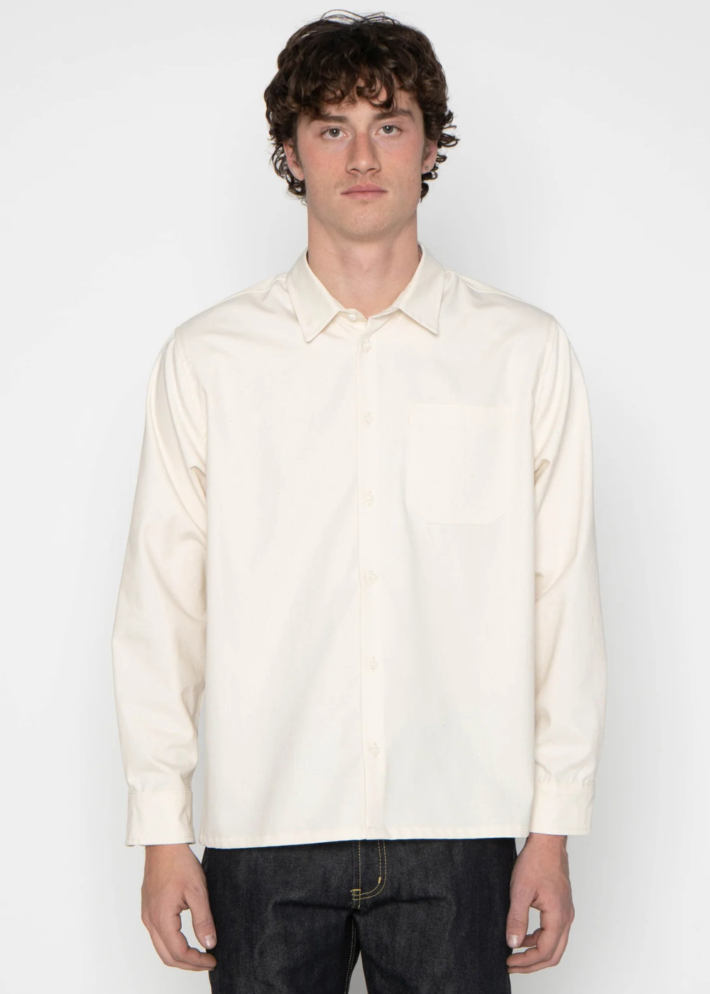 A man wearing a cream-colored long-sleeve button-down shirt and dark jeans, standing against a plain white studio background.