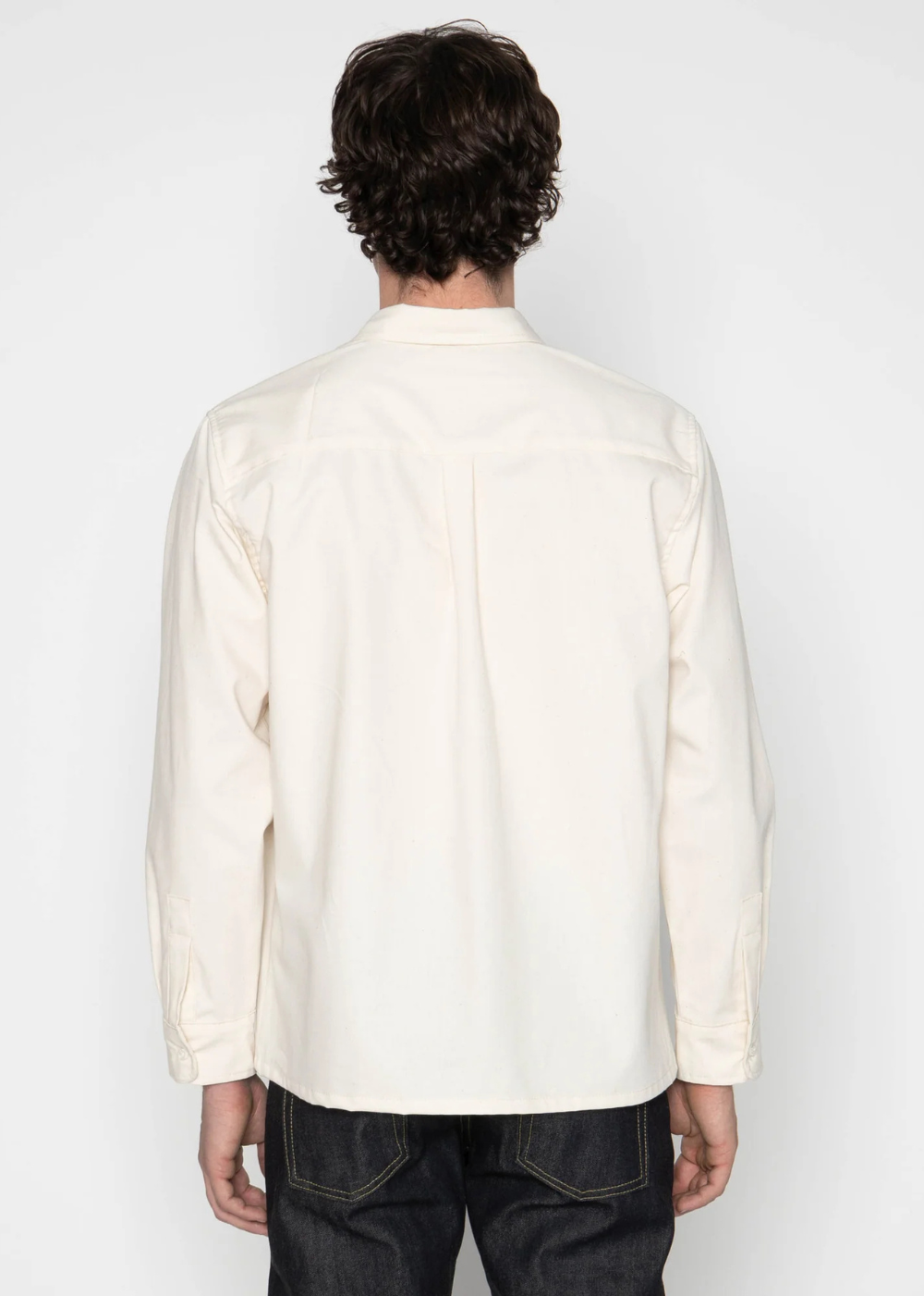 A man viewed from behind wearing a cream-colored long-sleeve button-up shirt and dark denim jeans, standing against a plain light gray studio background.