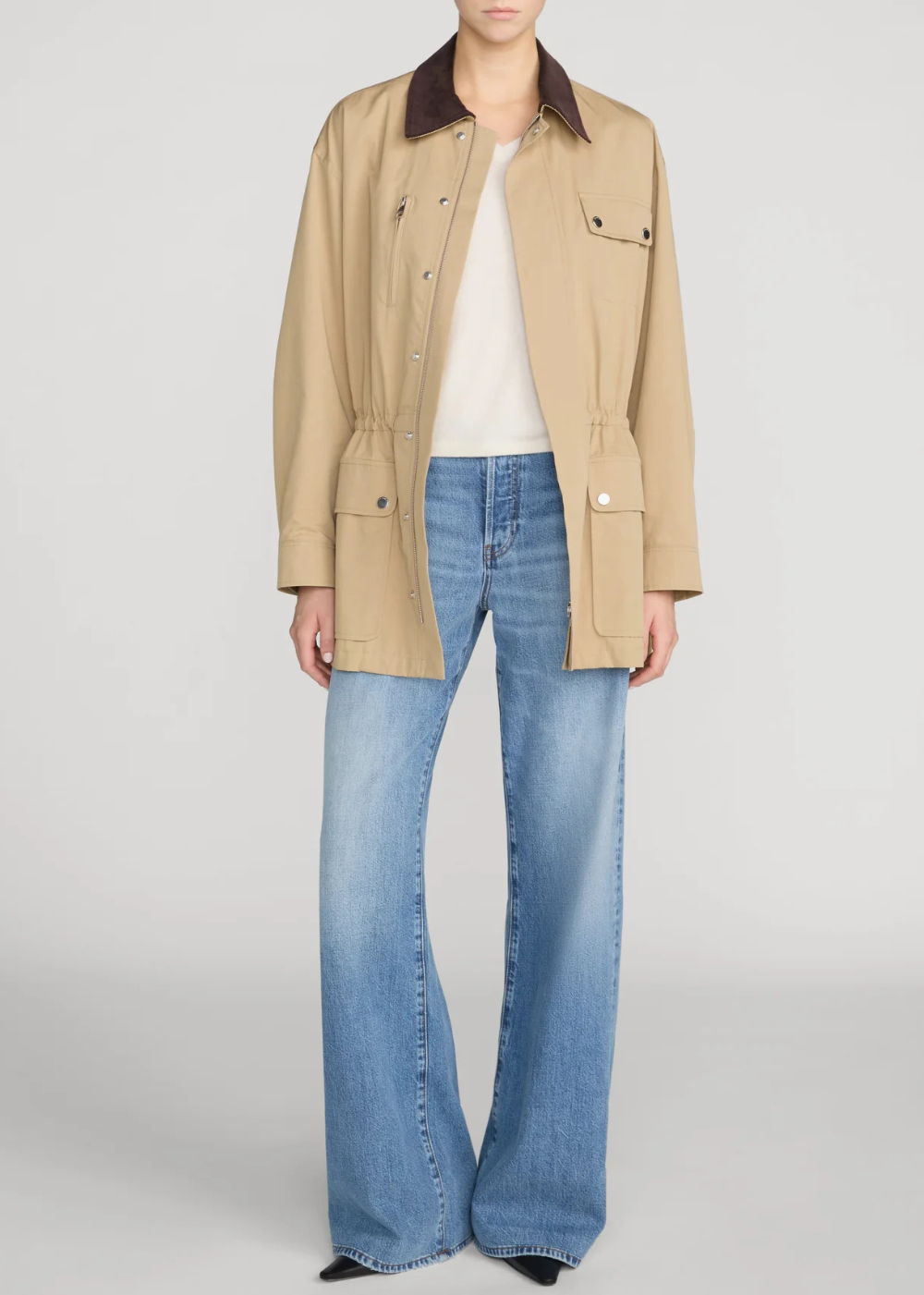 A model wearing a beige utility jacket with a dark brown collar, a white top, and light-wash blue flare jeans, photographed against a light gray studio background.