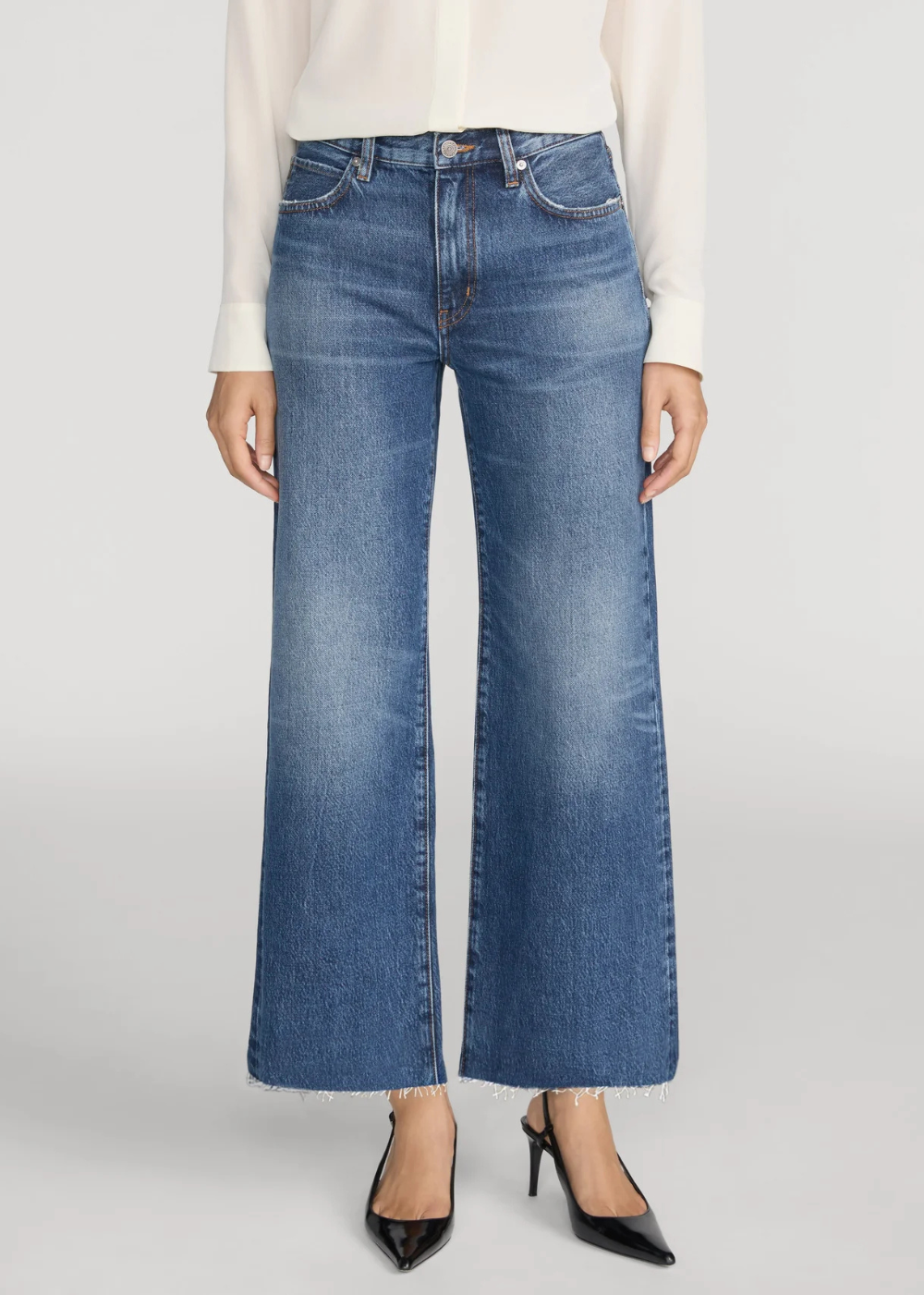 A model wearing a pair of medium-wash blue wide-leg cropped jeans with a faded wash, styled with a white blouse and black pointed-toe heels against a light gray studio background.