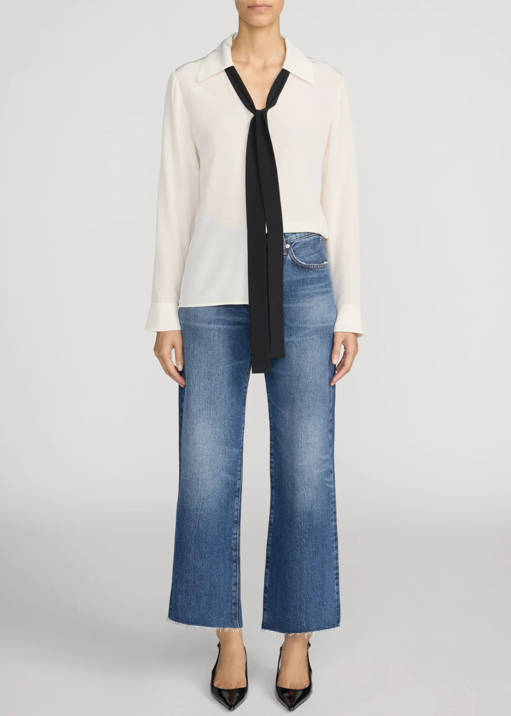 A model wearing a white long-sleeve button-up shirt with a black tie-neck scarf, blue wide-leg jeans, and black pointed-toe heels, photographed against a light gray studio background.