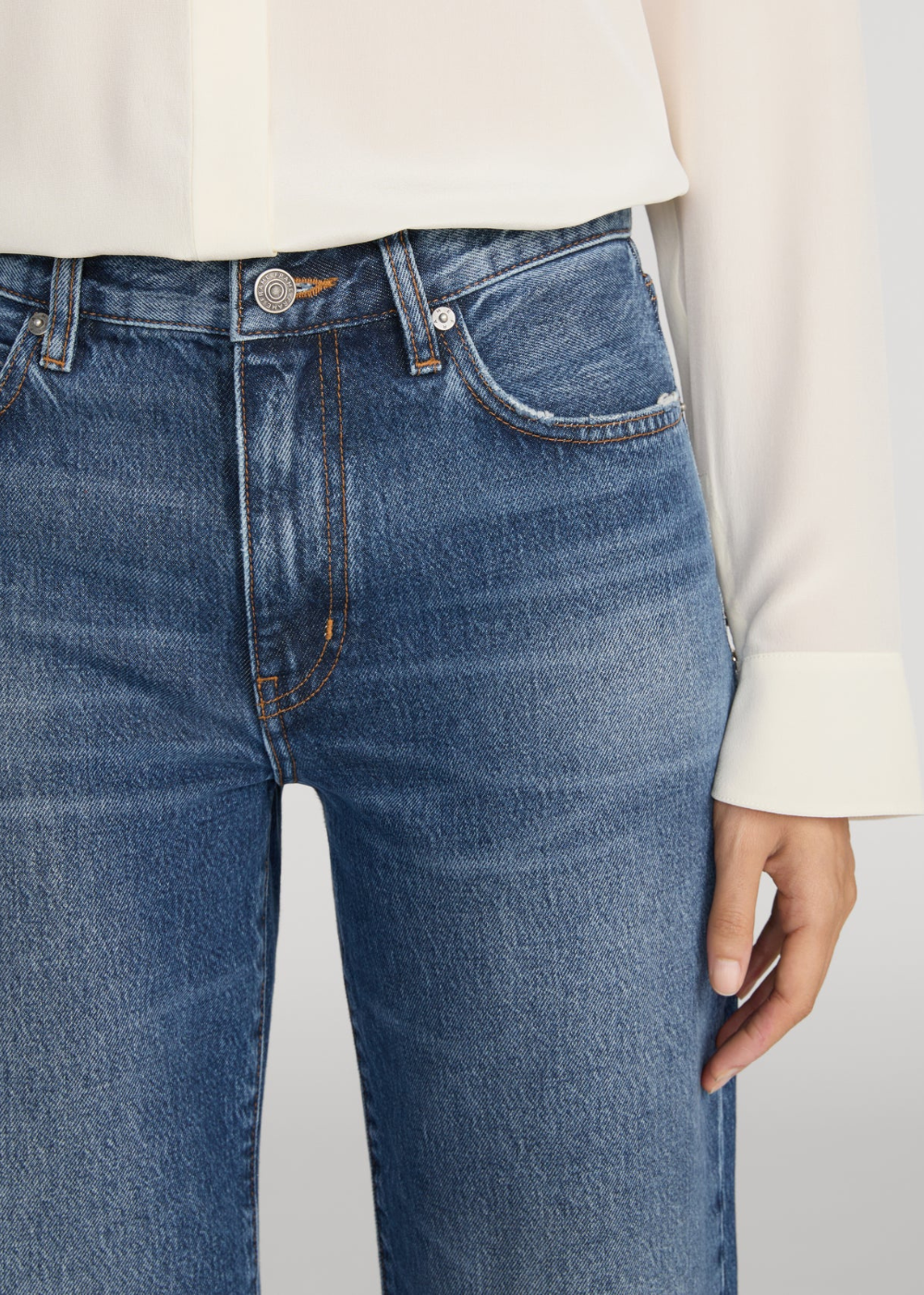 Close-up of a person wearing medium-wash blue denim jeans with a cream-colored blouse, showing the front waistband, pocket, and hand.