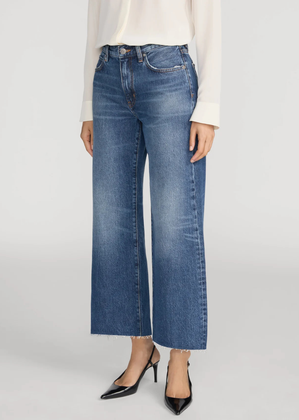 A model is wearing a pair of high-waisted, wide-leg cropped blue jeans with a frayed hem, styled with a cream long-sleeve top and black pointed-toe heels against a light gray studio background.