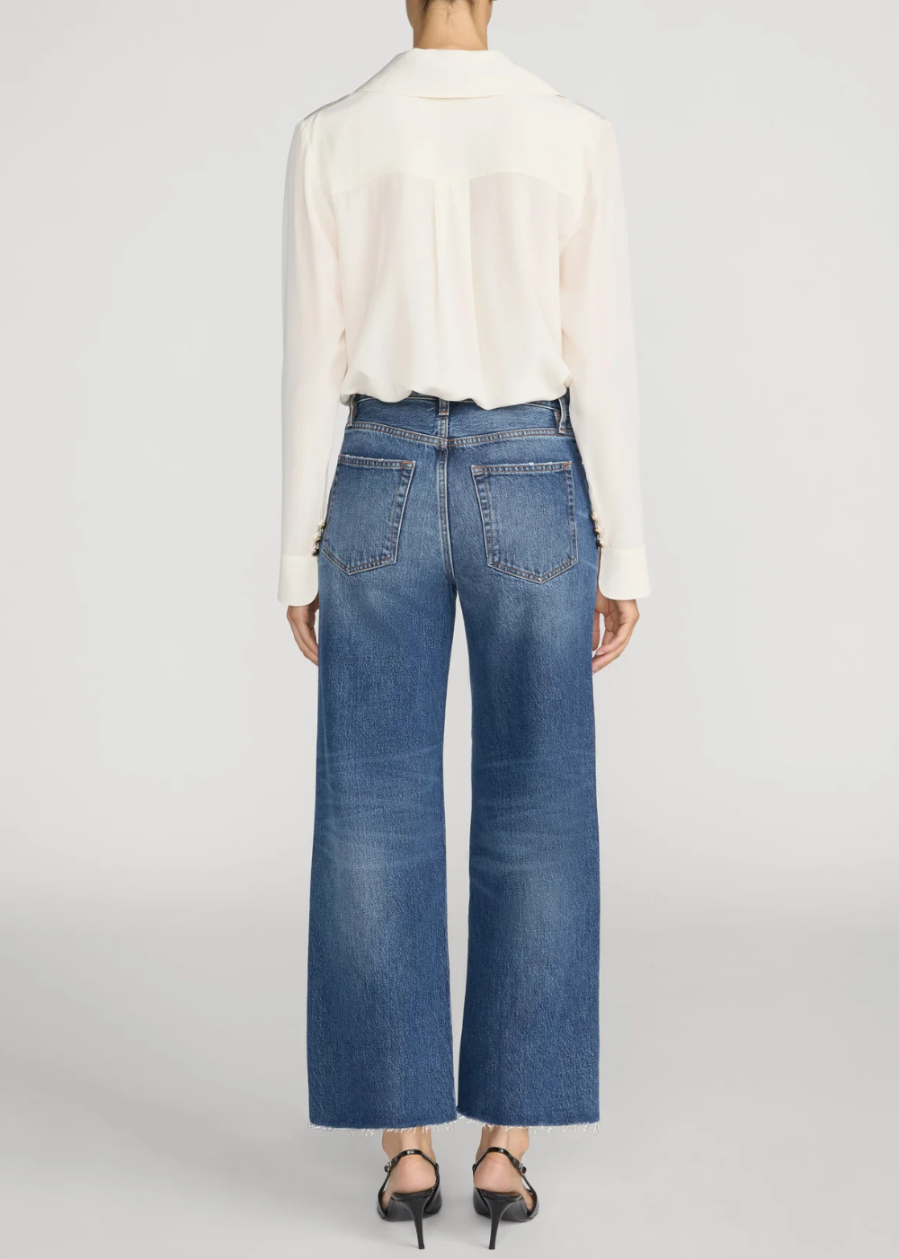 A model shown from the back wearing a cream long-sleeve blouse and blue wide-leg cropped jeans with black heels, against a light gray studio background.