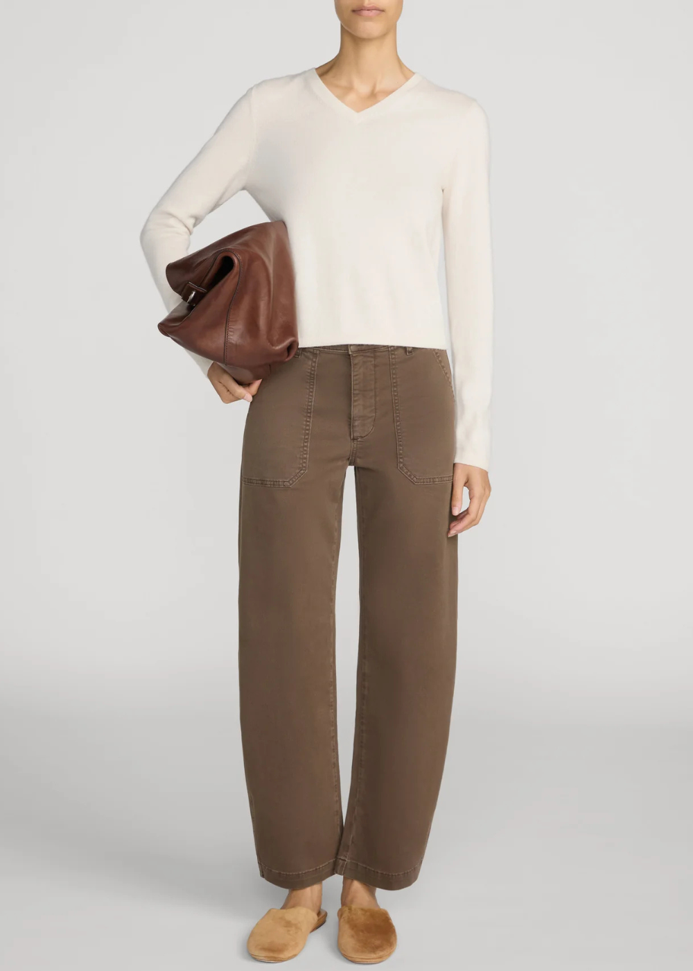 A model wearing a cream V-neck knit sweater, brown wide-leg pants, and tan slip-on shoes, holding a brown leather clutch against a light gray studio background.