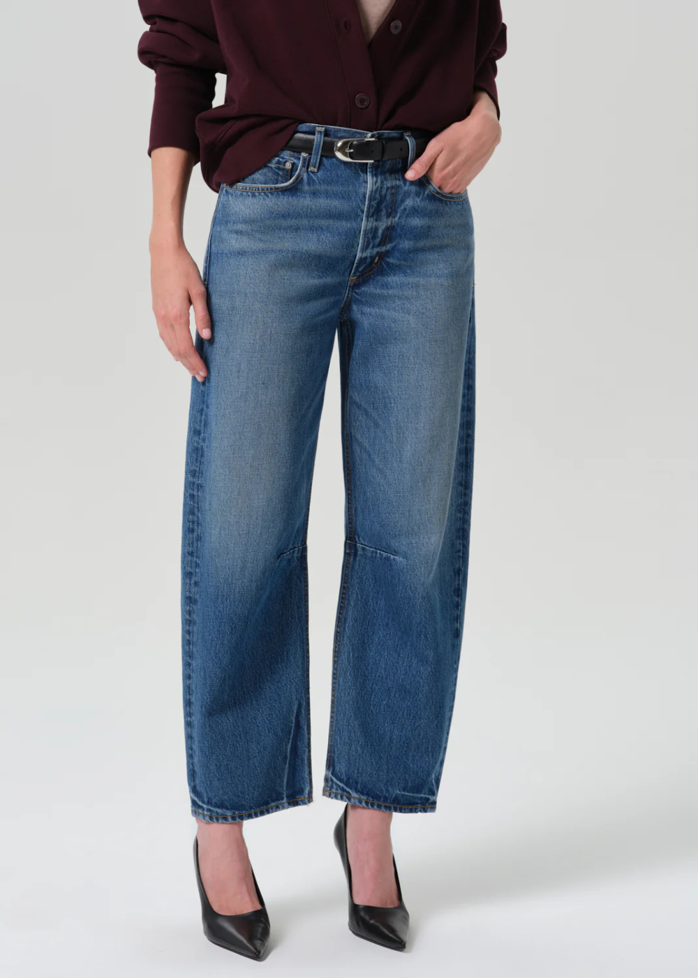 A model wearing a pair of wide-leg cropped blue denim jeans with a black belt, paired with a dark burgundy button-up shirt and black pointed-toe heels, against a light gray studio background.