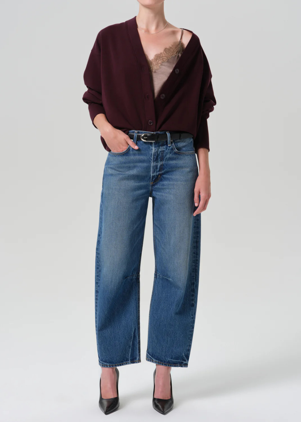 A model wearing a dark burgundy cardigan, blue wide-leg cropped jeans, a black belt, and black pointed-toe heels, photographed against a light gray studio background.