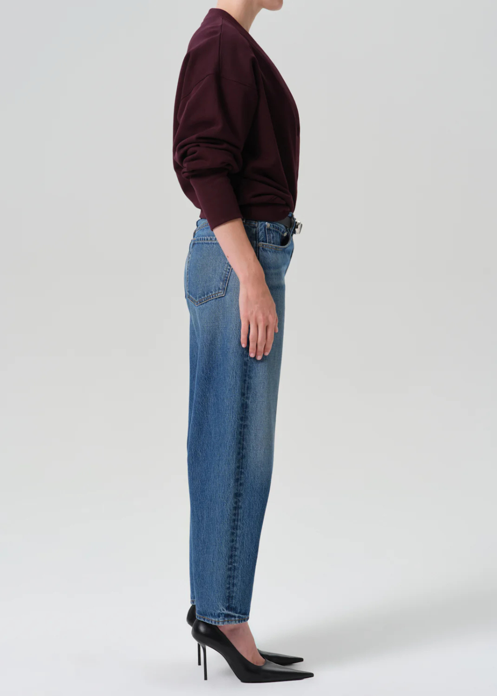 A side-profile view of a model wearing a dark burgundy long-sleeve top, blue wide-leg jeans, and black pointed-toe high heels against a light gray studio background.