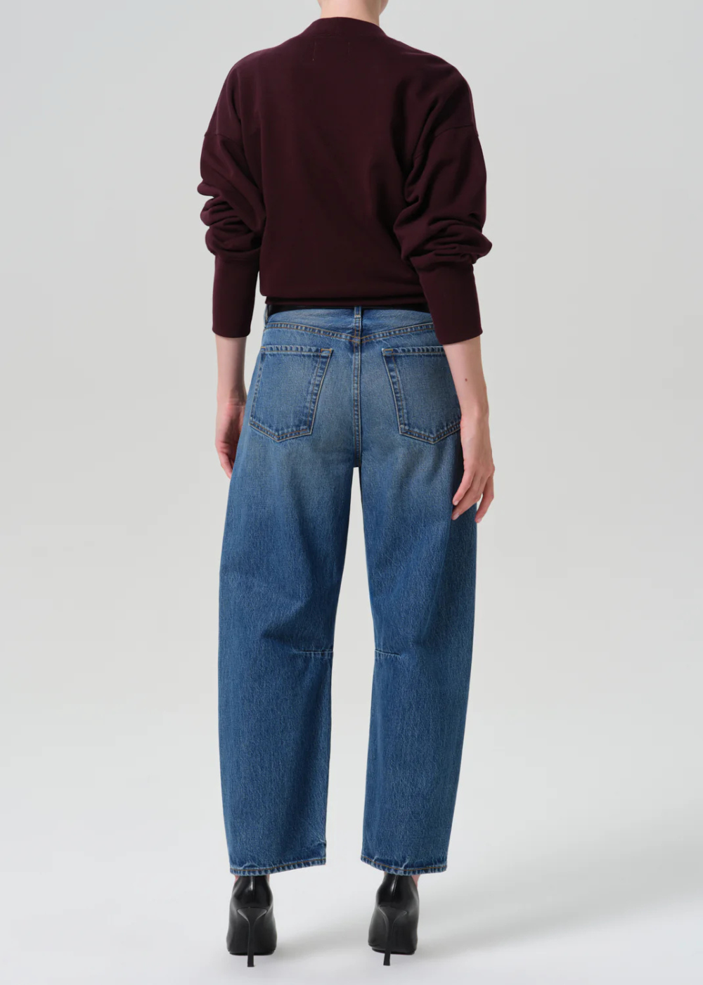 Rear view of a model wearing blue wide-leg jeans, a dark burgundy long-sleeve top, and black pointed-toe heels against a light gray studio background.
