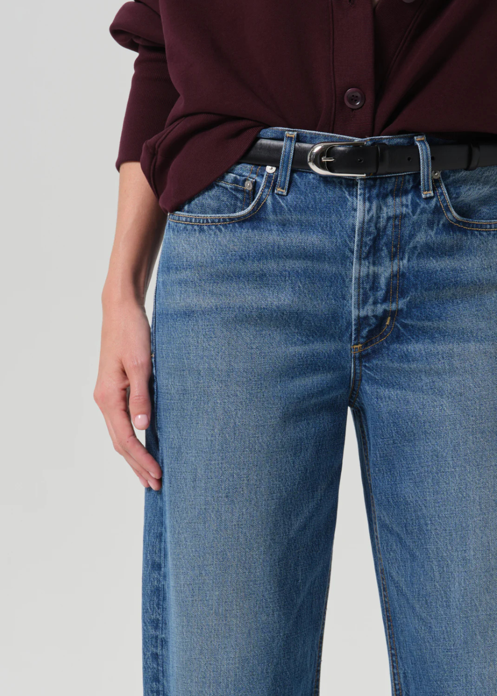 Close-up of a person wearing blue denim jeans with a black belt and a dark burgundy button-up shirt, against a light gray studio background.