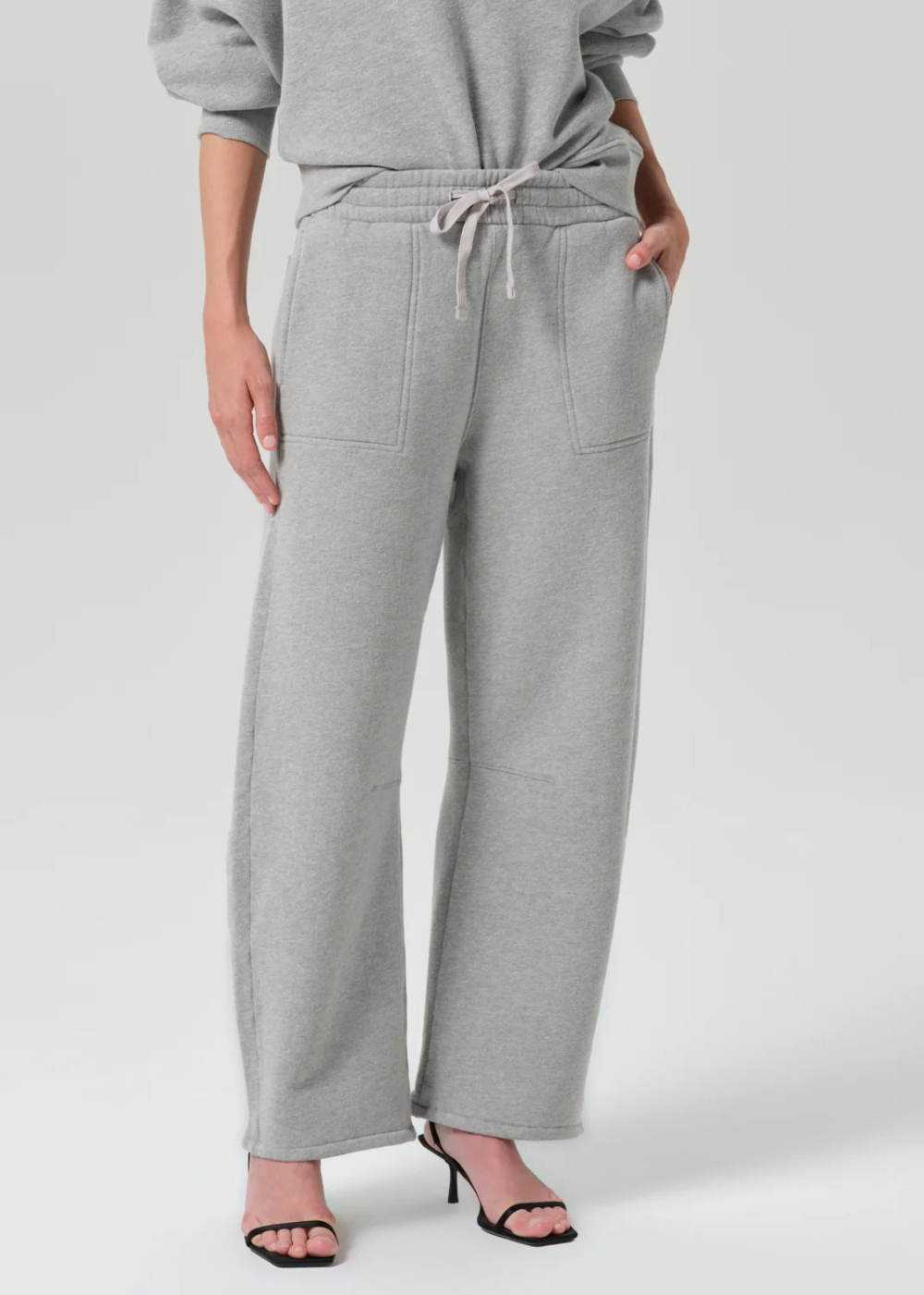 A person wearing light heather gray wide-leg sweatpants with a drawstring waistband, paired with a matching gray sweatshirt and black heeled sandals, photographed against a clean light gray studio background.