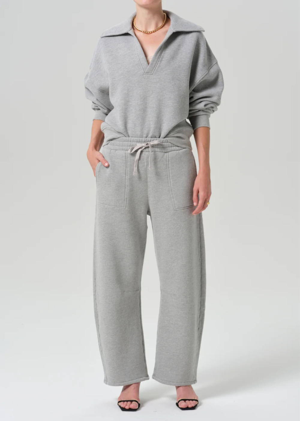 A model wearing a light gray two-piece loungewear set consisting of a half-zip sweatshirt and matching wide-leg sweatpants, photographed against a neutral light gray studio background.