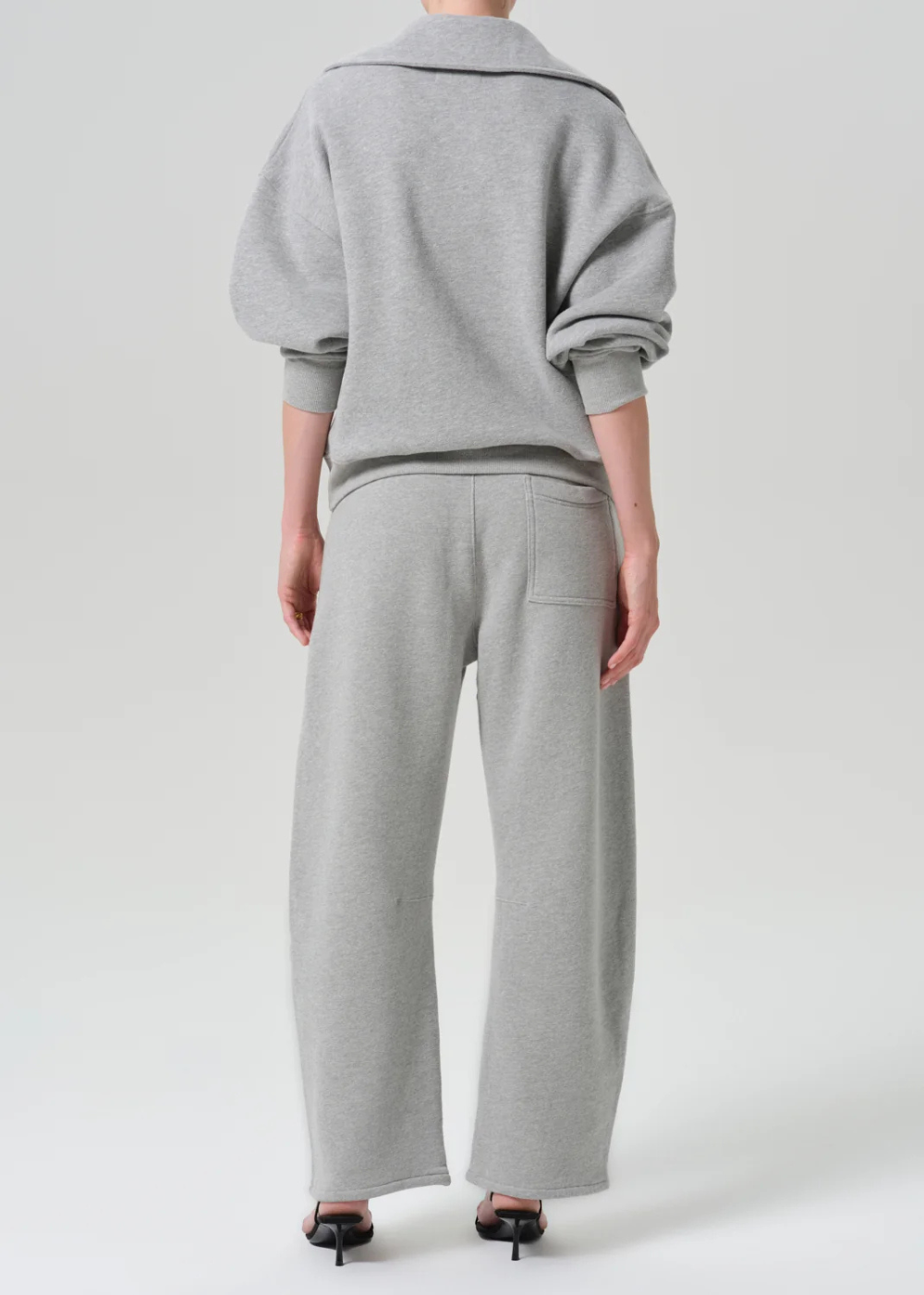 A model viewed from the back wearing a light gray heathered sweatshirt with voluminous sleeves and matching wide-leg sweatpants, paired with black high-heeled sandals against a light gray studio background.