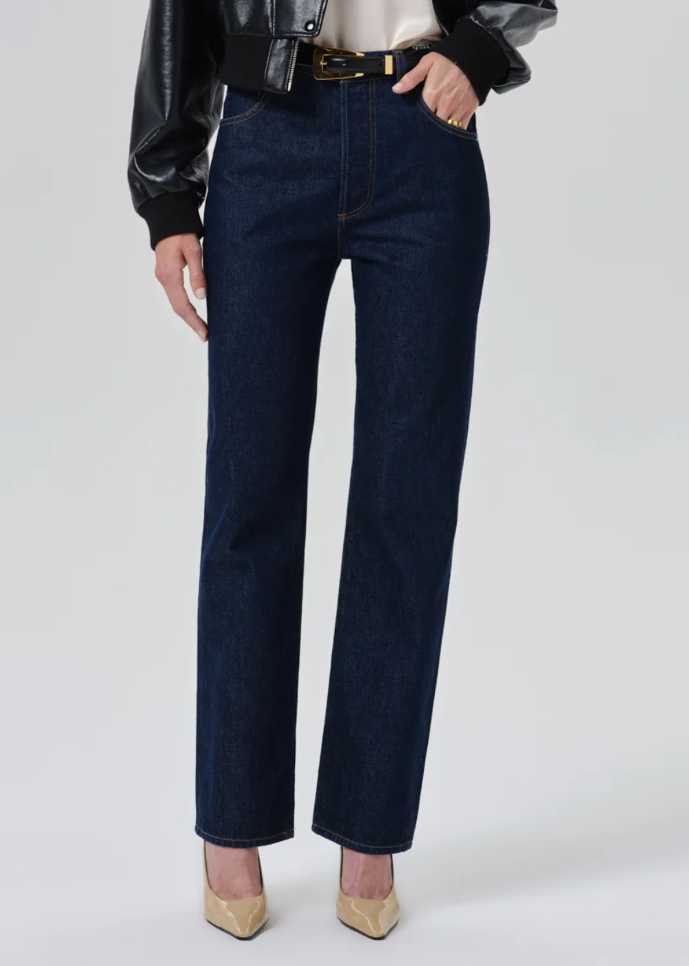A model wearing dark blue straight-leg denim jeans with a black leather jacket, white top, and beige heels, shown from the waist down on a light gray studio background.