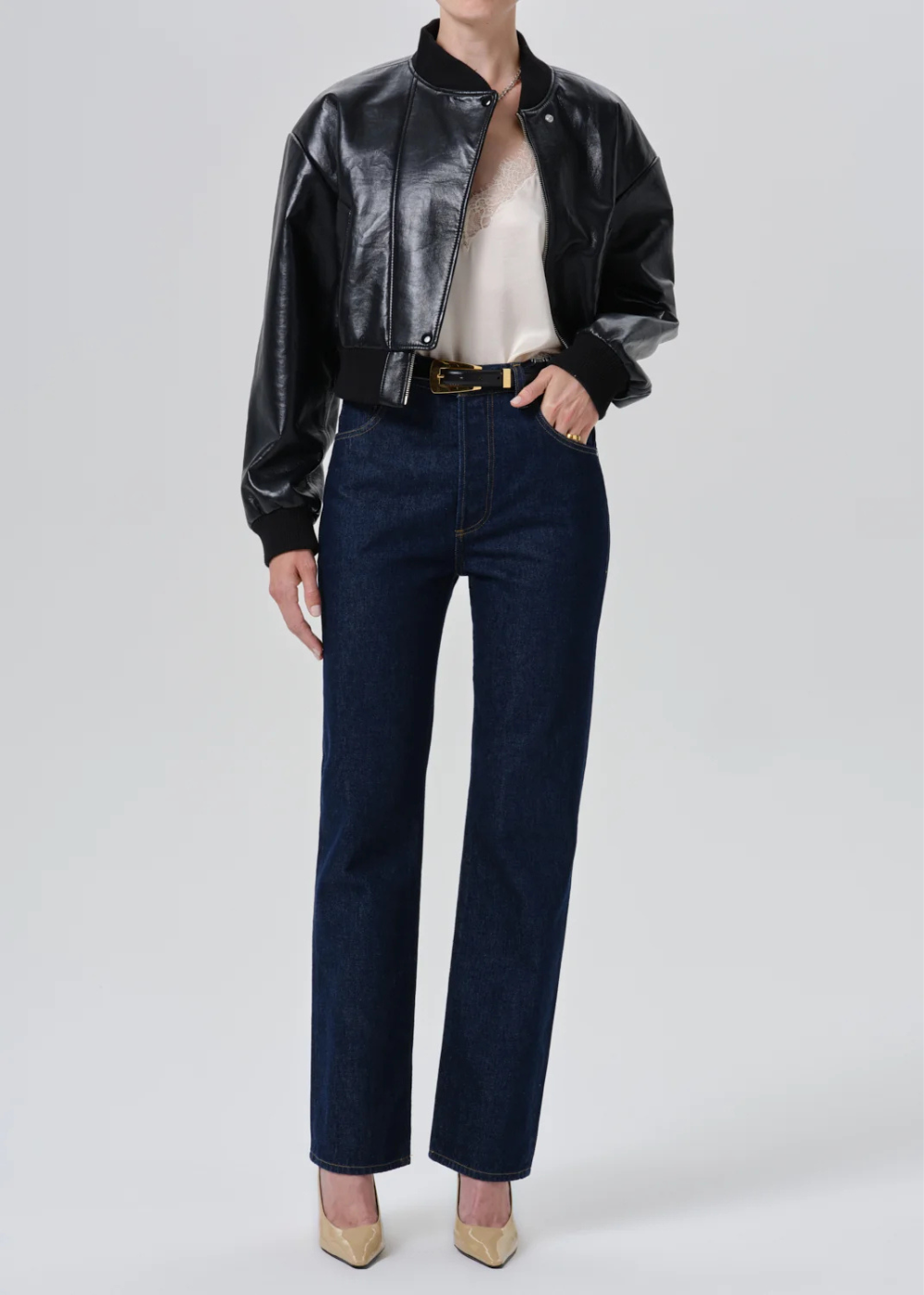 A model wearing a black leather bomber jacket, dark blue straight-leg jeans, a cream top, and beige heels, standing against a light gray studio background.