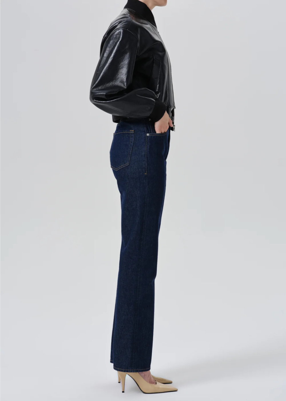 A side-profile studio shot of a model wearing dark blue straight-leg jeans, a black faux-leather jacket, and beige heels against a light gray background.