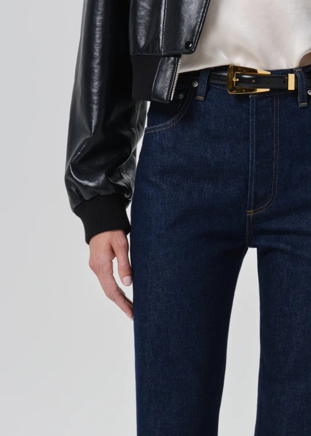 Close-up of a person wearing dark blue denim jeans with a black belt featuring a gold buckle, styled with a black faux-leather jacket and a cream top against a light gray background.