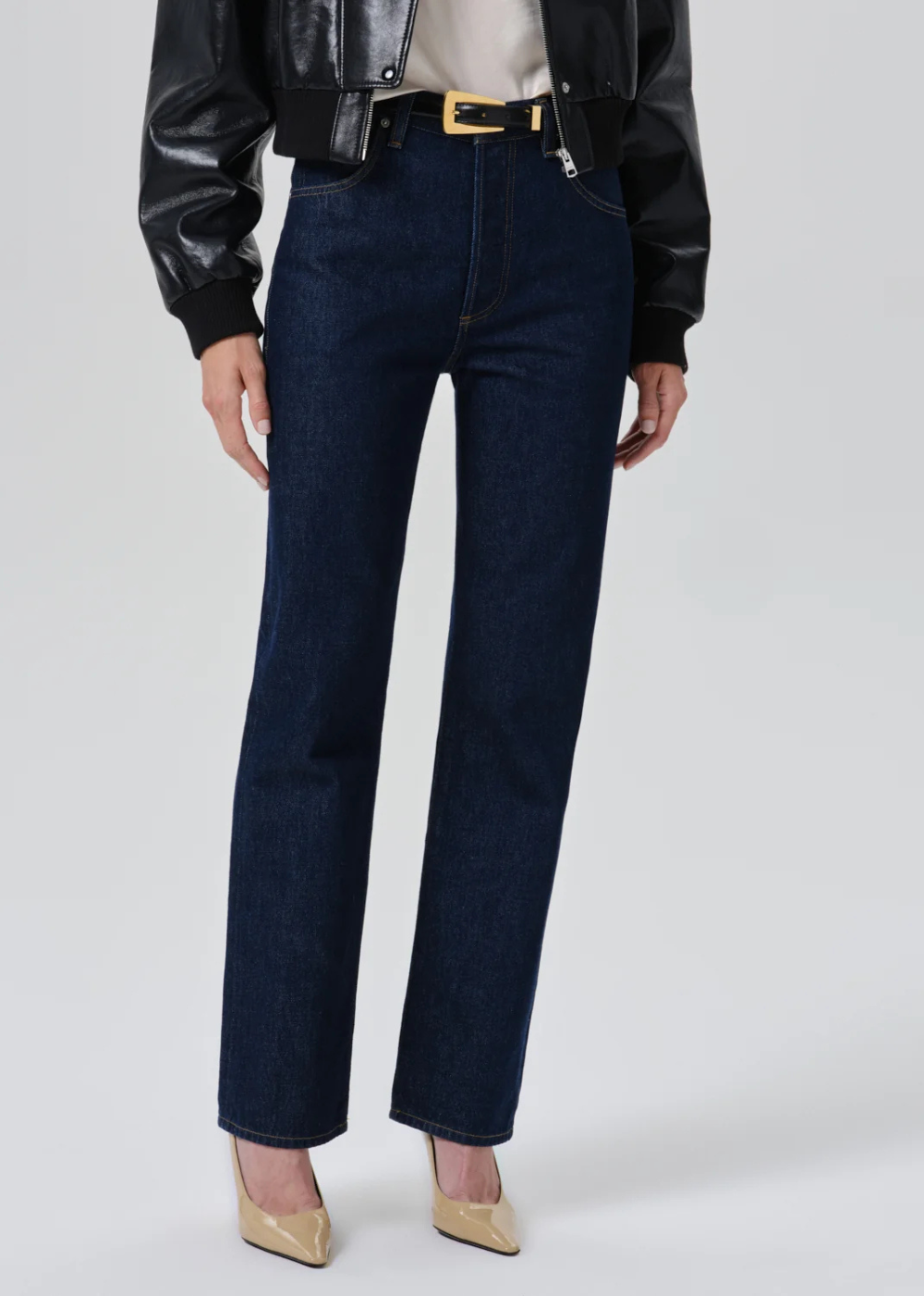 A model wearing dark blue straight-leg denim jeans with a gold-tone belt, styled with a black leather jacket, beige top, and beige pointed-toe heels against a light gray studio background.