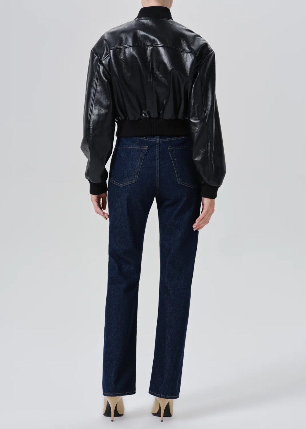 A model shown from the back wearing a black leather bomber jacket and dark blue straight-leg jeans, paired with beige high heels, against a light gray studio background.