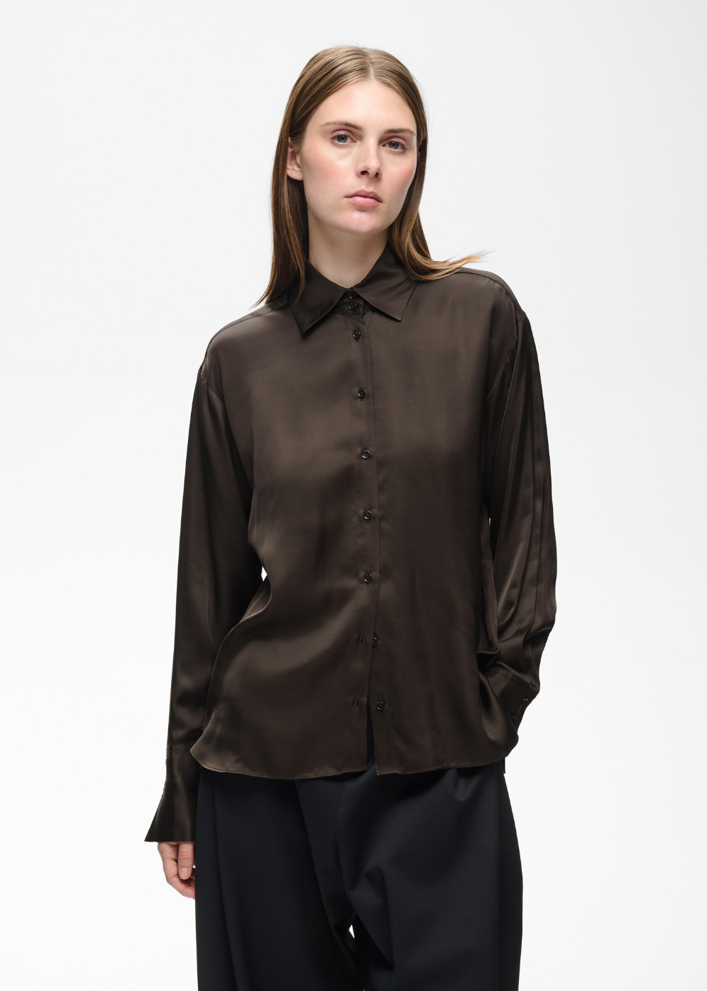 A woman modeling a dark brown long-sleeve button-up shirt with a satin-like sheen, paired with black trousers, against a plain white studio background.