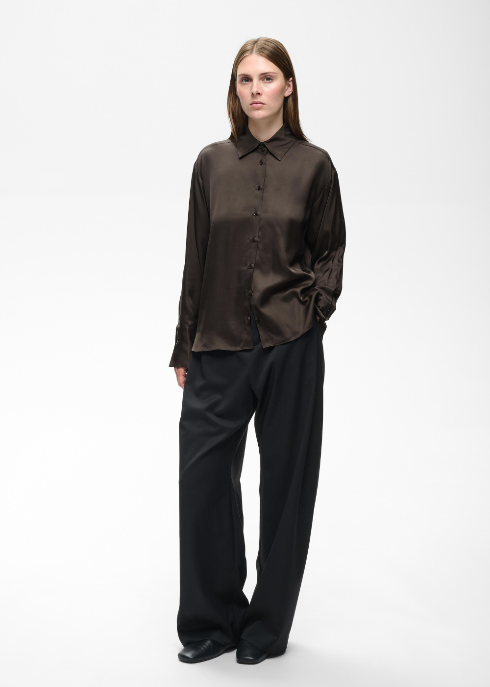 A model stands against a plain light gray studio background wearing a dark brown satin button-up shirt and loose black trousers.