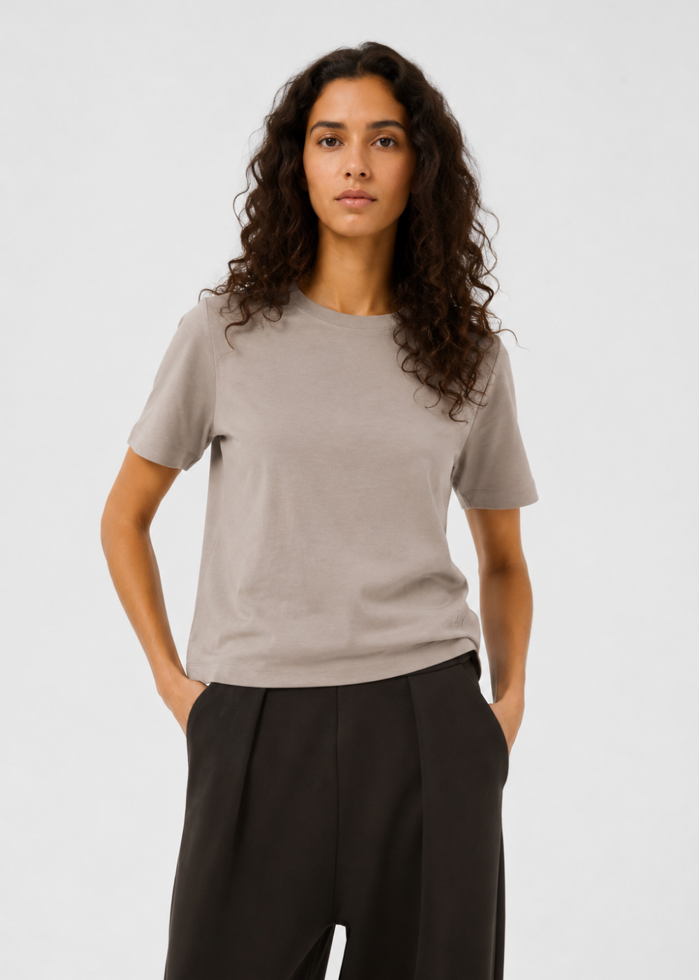 A woman with long, curly dark brown hair wearing a light taupe-gray short-sleeve crewneck t-shirt and black wide-leg trousers, standing against a light gray studio background.