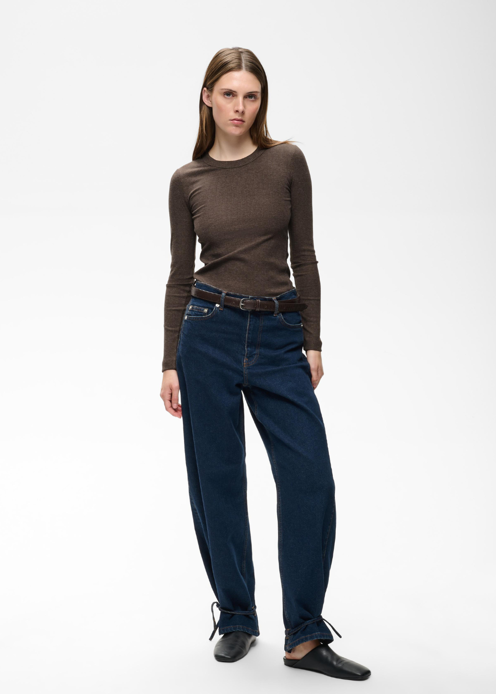 A woman modeling a brown long-sleeve knit top and dark blue straight-leg jeans with a brown belt, standing against a plain white studio background.