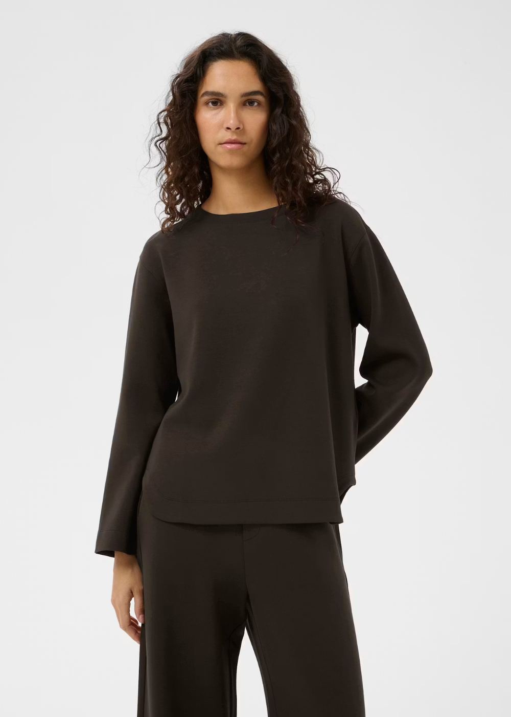 A woman with curly dark hair wearing a dark brown long-sleeve knit top and matching dark brown wide-leg pants, photographed against a light gray studio background.