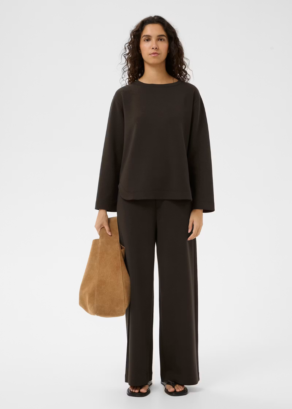 A woman stands against a light gray studio background wearing a dark brown long-sleeve top and matching wide-leg pants, holding a tan suede tote bag.