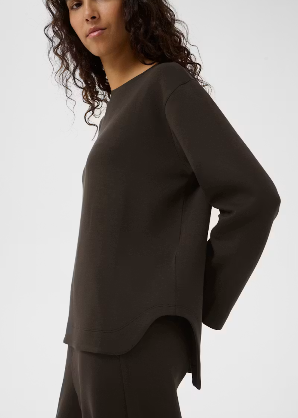 A woman wearing a dark brown long-sleeve knit top and matching dark brown bottoms, shown from the side against a light gray studio background.